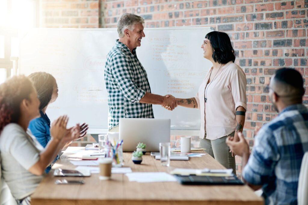 Ein Mann und eine Frau begrüßen sich per Handschlag im Büro eines Startups, lächeln und unterstützen die Einführung eines neuen Teammitglieds. Geschäftsleute und Führungskräfte schütteln dem neuen Mitarbeiter die Hand, um einen Deal, eine Vereinbarung und die erfolgreiche Integration ins Team zu feiern.