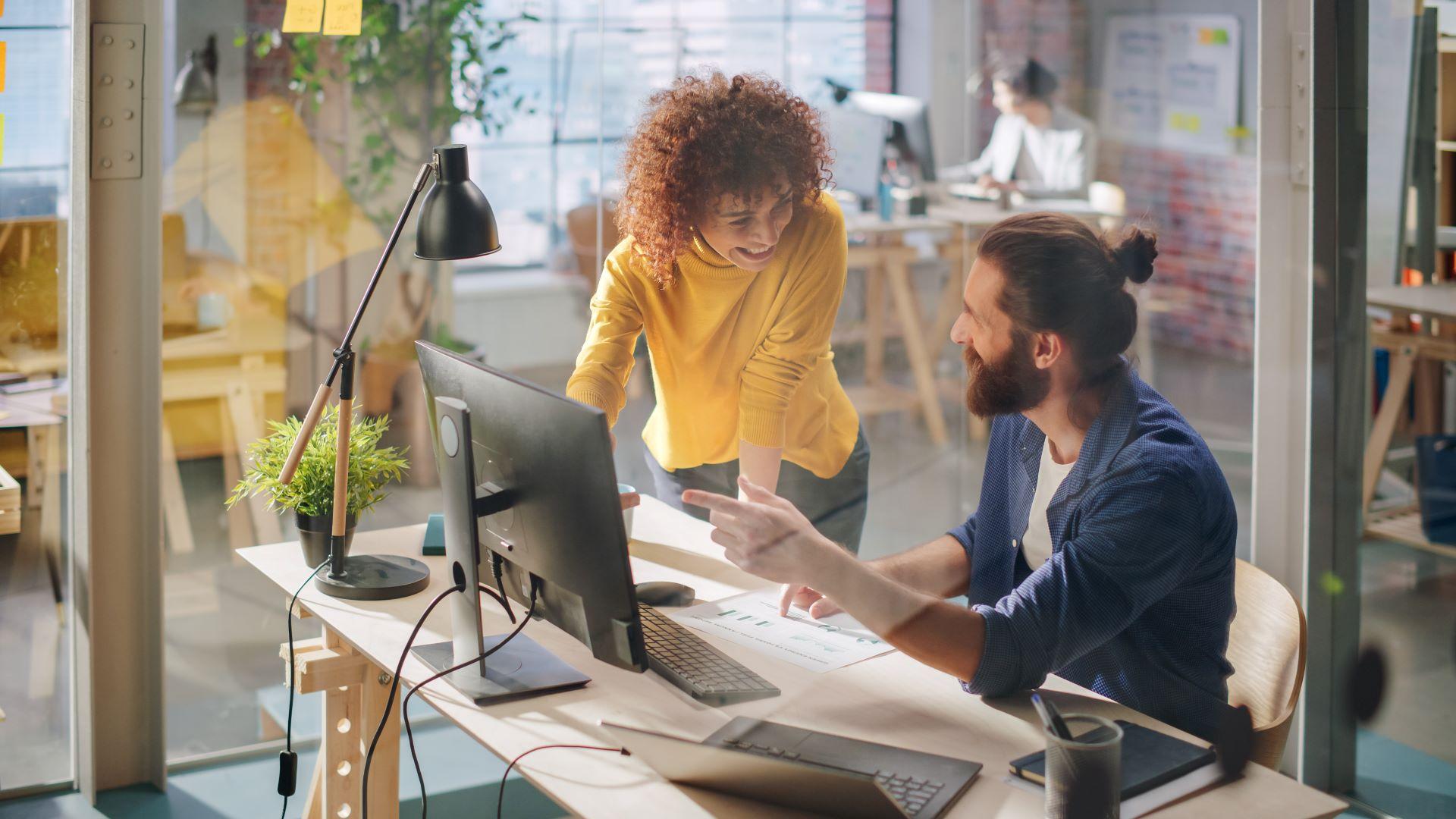 Zwei Mitarbeiter in einem modernen Büro, die gemeinsam an einem Computer arbeiten. Die Frau im gelben Pullover lehnt sich über den Tisch und lacht, während der Mann ihr etwas auf dem Bildschirm zeigt. Im Hintergrund sind weitere Arbeitsplätze und eine kreative Arbeitsumgebung zu sehen.