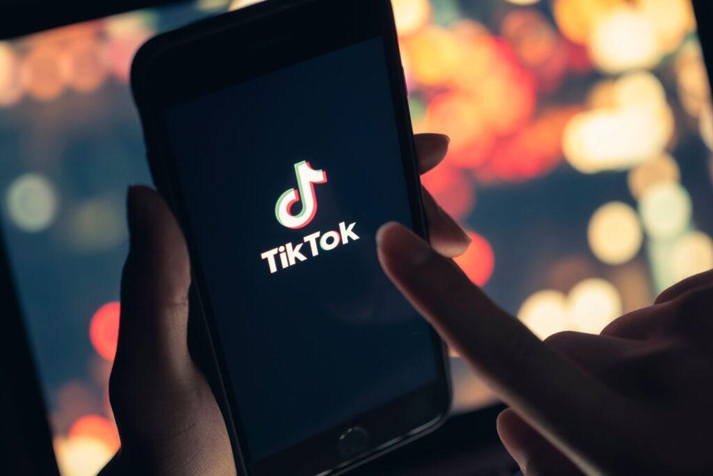 Nahaufnahme einer Hand, die ein Smartphone hält, auf dem das TikTok-Logo auf dem Bildschirm angezeigt wird. Der Hintergrund ist unscharf, zeigt aber bunte Lichter, die an eine Stadt bei Nacht erinnern.